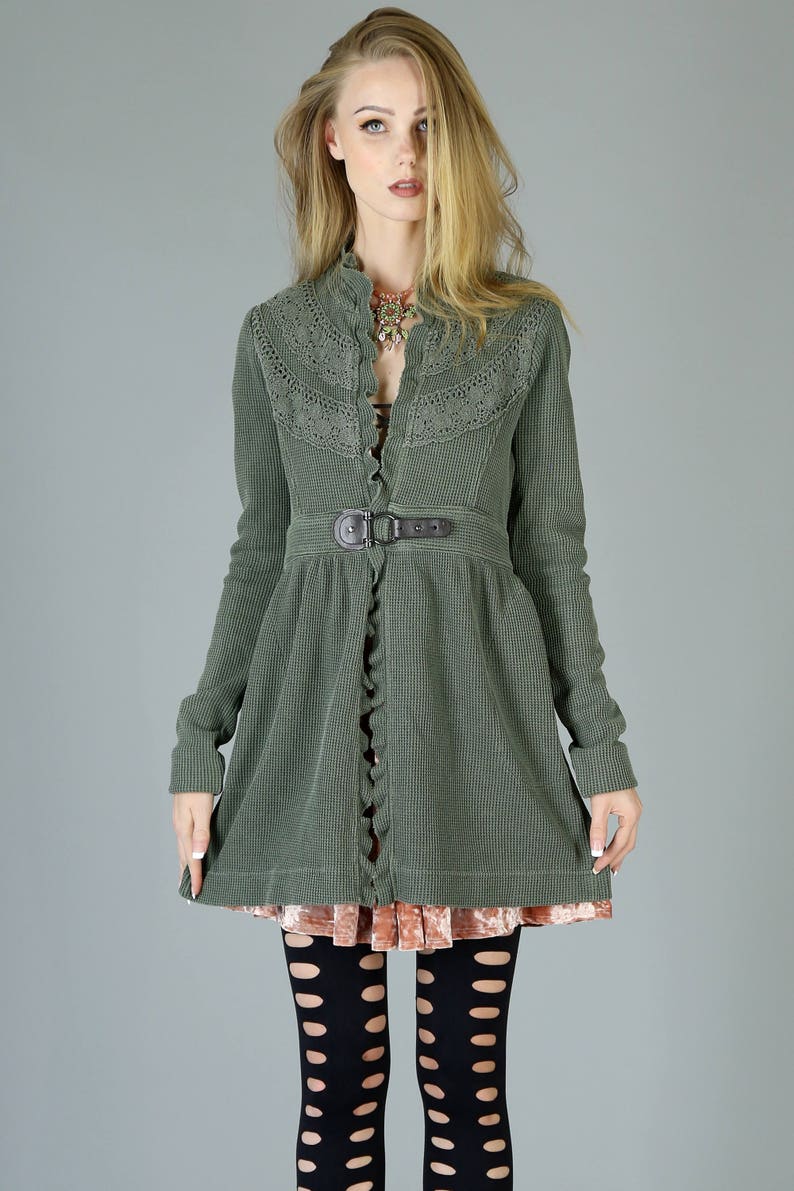 May include: A green waffle knit jacket with a lace trim, a brown belt, and a ruffled hem. The jacket is worn over a pink velvet skirt and black tights with cutouts.