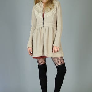 Op de afbeelding: Een beige gebreide cardigan met een kanten rand en een ritssluiting. De cardigan wordt gedragen over een rode jurk en zwarte kanten panty's. Het model draagt zwarte laarzen met hakken.