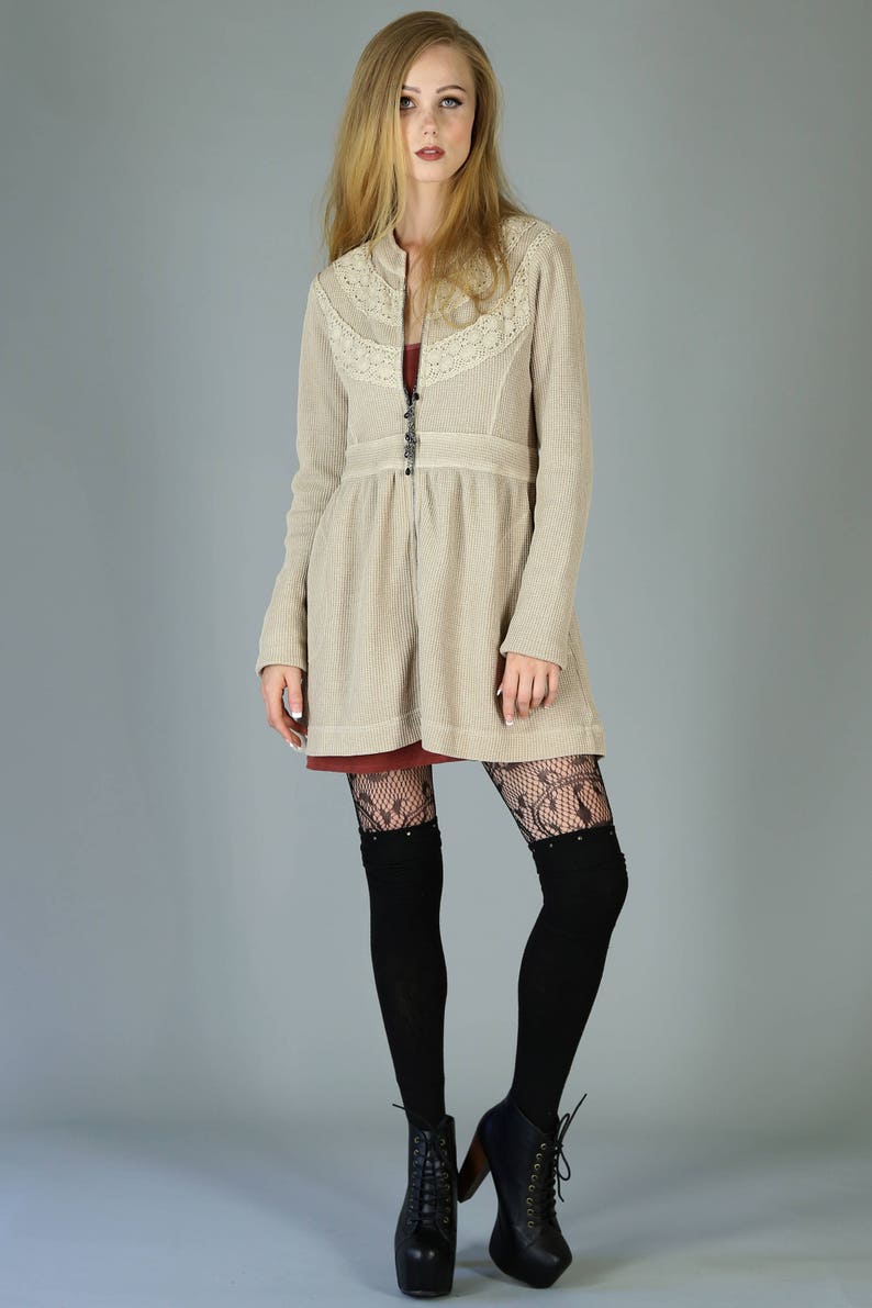 Op de afbeelding: Een beige gebreide cardigan met een kanten rand en een ritssluiting. De cardigan wordt gedragen over een rode jurk en zwarte kanten panty's. Het model draagt zwarte laarzen met hakken.