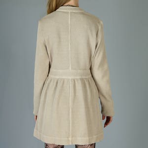 Op de afbeelding: Een beige wafel gebreide cardigan met lange mouwen en een getailleerde taille. De cardigan wordt gedragen over zwarte kanten panty's.