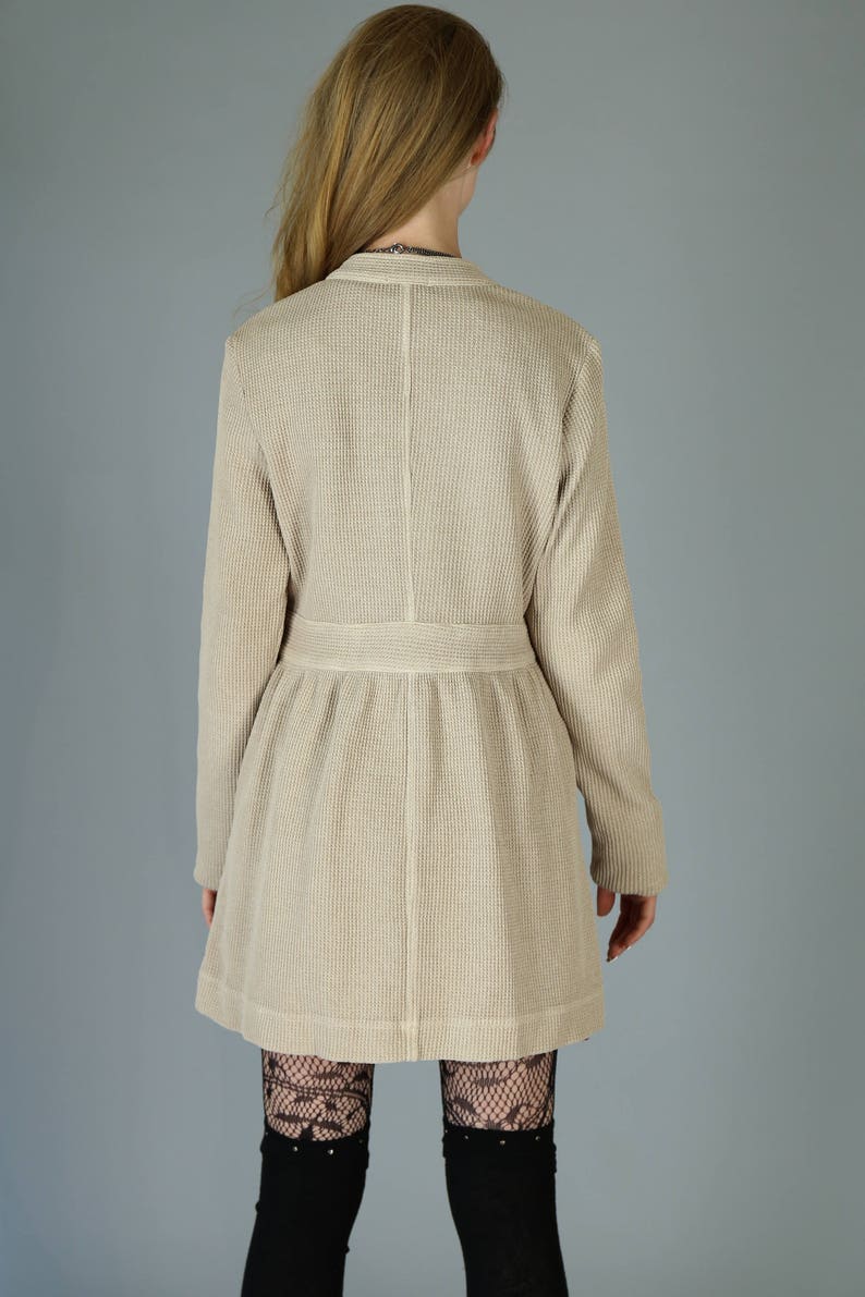 Op de afbeelding: Een beige wafel gebreide cardigan met lange mouwen en een getailleerde taille. De cardigan wordt gedragen over zwarte kanten panty's.