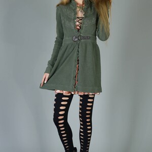 May include: A green cardigan with a lace overlay and a belt. The cardigan is worn over a pink dress and black tights with cutouts. The model is wearing black heeled boots.