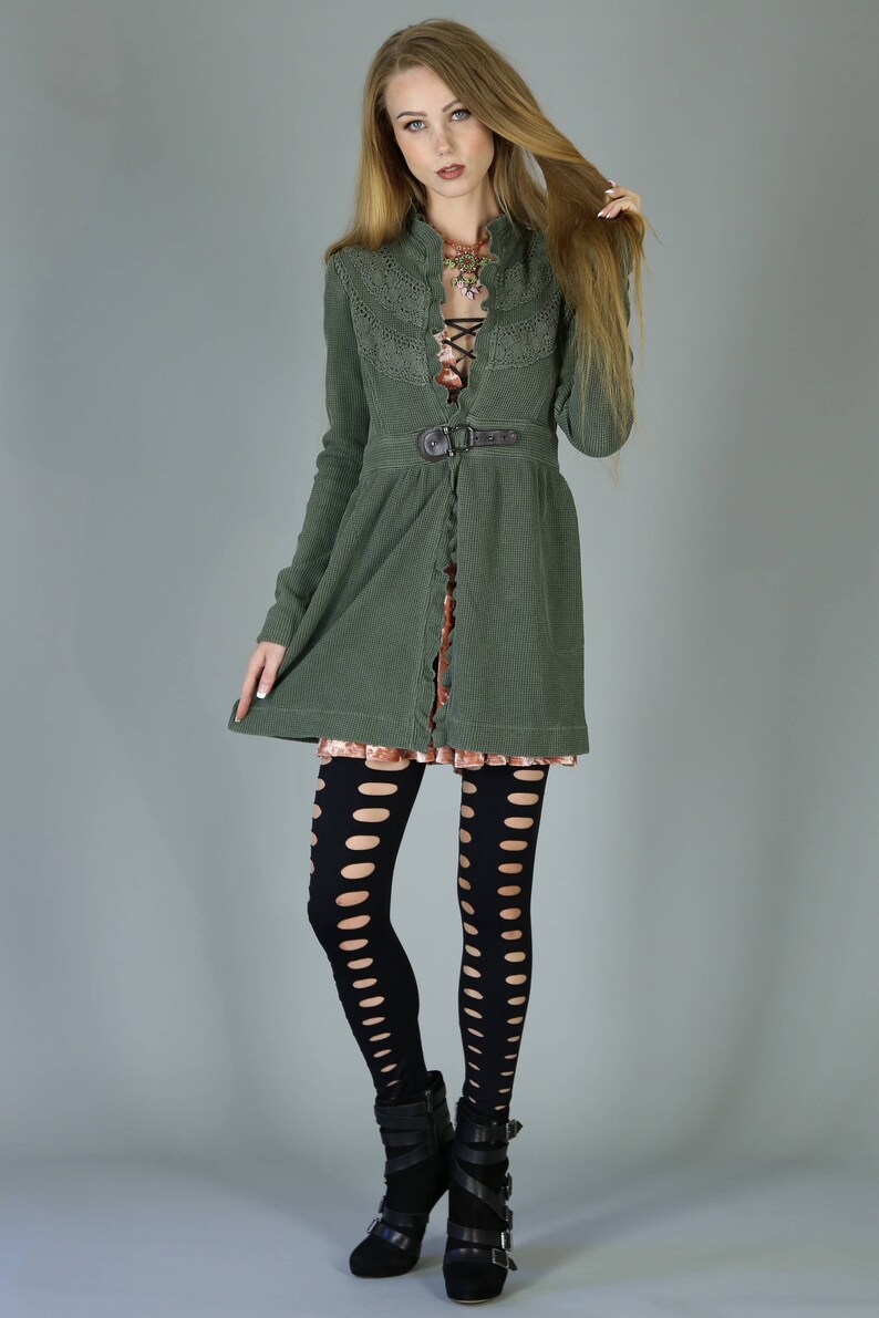 May include: A green cardigan with a lace overlay and a belt. The cardigan is worn over a pink dress and black tights with cutouts. The model is wearing black heeled boots.