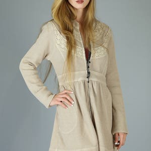 Op de afbeelding: Een beige wafel gebreide cardigan met een kantversiering en een ritssluiting. De cardigan heeft een getailleerde pasvorm en een uitlopende rok. Het model draagt zwarte kousen met een visnetpatroon.