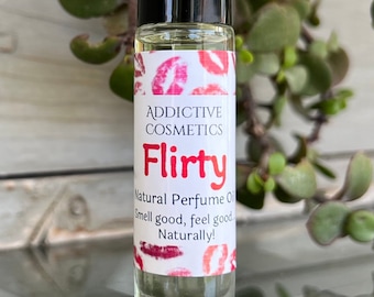 May include: A clear glass roll-on bottle with a black cap and a white label. The label says "Addictive Cosmetics" and "Flirty" in red letters. The label also says "Natural Perfume Oil" and "Smell good, feel good, Naturally!" in black letters. The bottle is surrounded by green leaves.