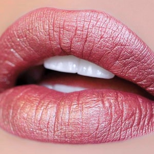 May include: Close-up of a person's lips, coated in a shimmering, metallic rose-colored lipstick. The lips are slightly parted, revealing bright white teeth. The lipstick has a textured appearance.