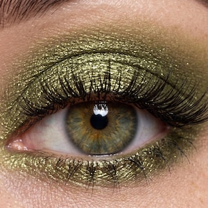May include: Close-up of an eye with green eyeshadow and long, black eyelashes. The eye is hazel with a bright highlight. The eyeshadow has a metallic, shimmering finish, creating a glamorous look.