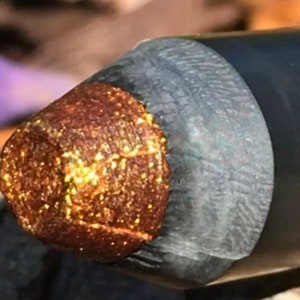May include: Close-up of a gold and brown eyeliner pencil with a metallic finish.