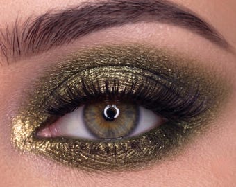 May include: Close-up of an eye with olive green eyeshadow and black mascara. The eye has a light brown iris with a dark ring around the pupil. The eyebrow is dark brown and well-groomed. The eyeshadow has a metallic shimmer.