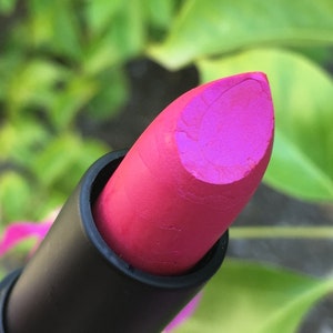 Pu&ograve; includere: Primo piano di un rossetto rosa vibrante. Il rossetto ha una finitura opaca ed &egrave; racchiuso in un tubetto nero. Lo sfondo &egrave; verde sfocato, che suggerisce un ambiente esterno.