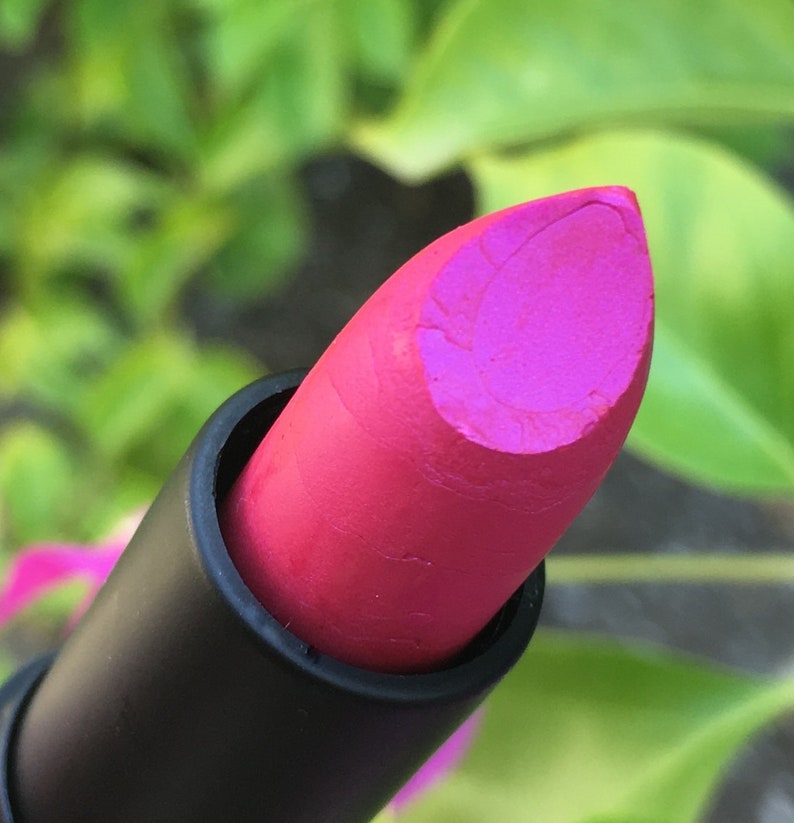 Pu&ograve; includere: Primo piano di un rossetto rosa vibrante. Il rossetto ha una finitura opaca ed &egrave; racchiuso in un tubetto nero. Lo sfondo &egrave; verde sfocato, che suggerisce un ambiente esterno.