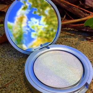 May include: A silver compact with a round, iridescent white powder inside. The compact is open and the mirror reflects a blurry image of a blue sky and green trees.