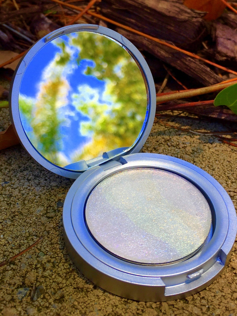 May include: A silver compact with a round, iridescent white powder inside. The compact is open and the mirror reflects a blurry image of a blue sky and green trees.