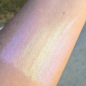 May include: A close-up of iridescent highlighter on skin, showing a pearly white, light pink, and light yellow shimmer.