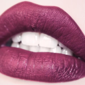 May include: Close-up of a mouth with deep magenta lipstick. The lips are full and textured, with a matte finish. The teeth are bright white, contrasting with the dark lipstick. The image is well-lit, highlighting the details of the lips.