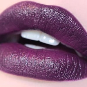 Puede incluir: Primer plano de una boca con labial morado oscuro. Los labios son carnosos y brillantes, con una forma definida. Los dientes blancos son visibles entre los labios. El labial tiene una textura rica y aterciopelada.