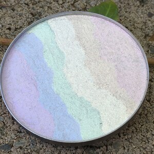 May include: A round, silver compact with a swirl of pastel colors, including pink, purple, blue, and green.