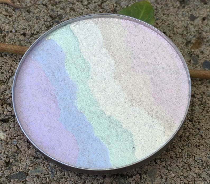 May include: A round, silver compact with a swirl of pastel colors, including pink, purple, blue, and green.