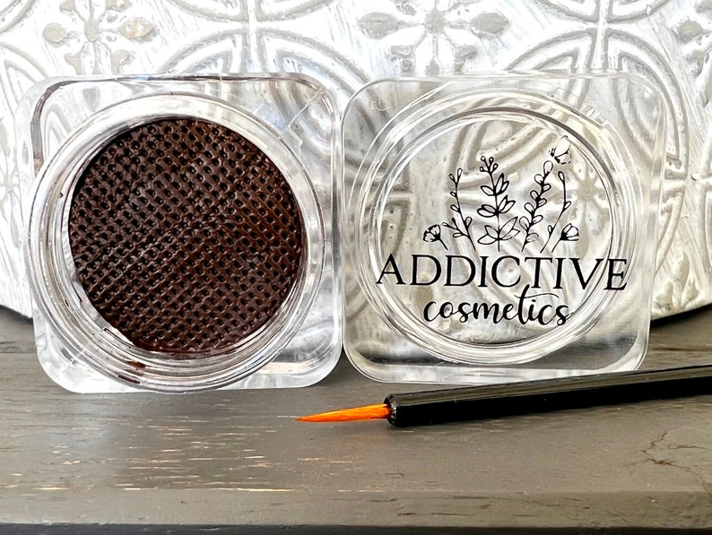 Pode incluir: Um pequeno recipiente de pl&aacute;stico transparente com um produto circular marrom texturizado dentro. O recipiente tem um r&oacute;tulo preto com o texto "ADDICTIVE cosmetics" e um design floral. Um pequeno pincel preto e laranja est&aacute; na frente do recipiente.