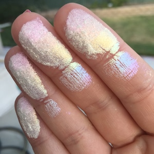 May include: Five fingers with swatches of iridescent highlighter makeup. The swatches are in shades of white, pink, and yellow with a rainbow sheen.