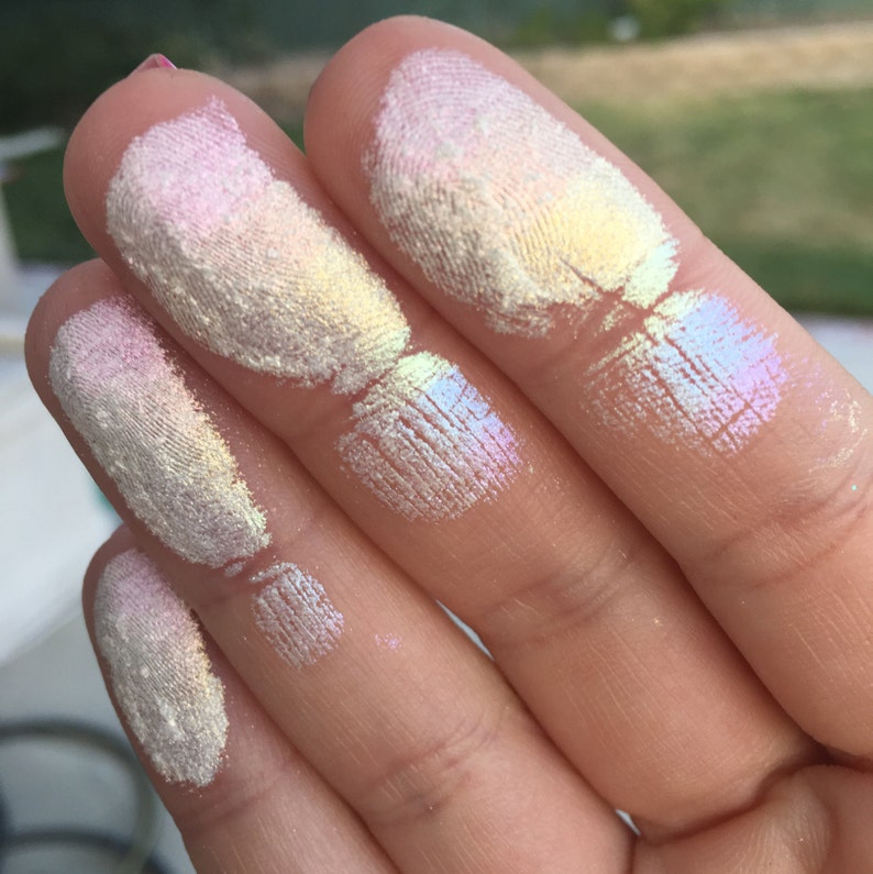 May include: Five fingers with swatches of iridescent highlighter makeup. The swatches are in shades of white, pink, and yellow with a rainbow sheen.