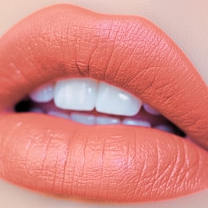 May include: Close-up of a mouth with coral-colored lipstick. The lips are full and well-defined, with a matte finish. The teeth are white and straight. The overall effect is a vibrant and polished look, perfect for beauty and makeup listings.