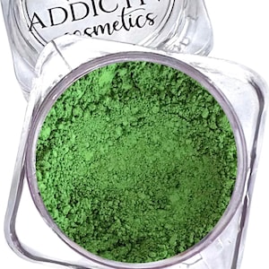 Puede incluir: Un tarro cosm&eacute;tico cuadrado transparente lleno de polvo verde brillante. El tarro est&aacute; parcialmente abierto, revelando el pigmento finamente molido. El nombre de la marca "ADDICTIVE cosmetics" es visible en la tapa.