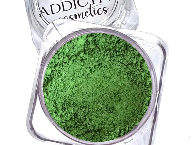 Puede incluir: Un tarro cosm&eacute;tico cuadrado transparente lleno de polvo verde brillante. El tarro est&aacute; parcialmente abierto, revelando el pigmento finamente molido. El nombre de la marca "ADDICTIVE cosmetics" es visible en la tapa.