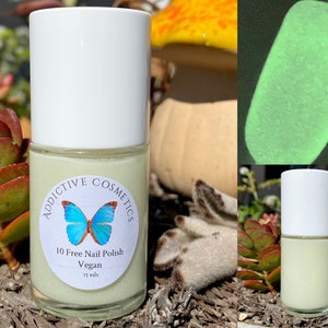 May include: A small bottle of light green nail polish with a white label that says "Addictive Cosmetics 10 Free Nail Polish Vegan 15 mls" and a blue butterfly graphic. The bottle is sitting on a bed of brown mulch and green plants.