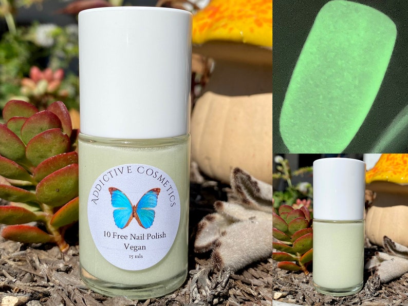 May include: A small bottle of light green nail polish with a white label that says "Addictive Cosmetics 10 Free Nail Polish Vegan 15 mls" and a blue butterfly graphic. The bottle is sitting on a bed of brown mulch and green plants.