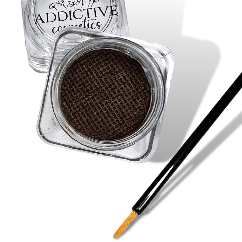 Pode incluir: Um pequeno recipiente quadrado transparente com um delineador de olhos l&iacute;quido castanho no seu interior. O recipiente tem o texto "ADDICTIVE cosmetics" na parte superior. Tamb&eacute;m est&aacute; inclu&iacute;do um pincel delineador de olhos preto com ponta dourada.