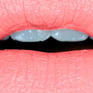 May include: Close-up of a person's lips, painted with a coral-colored lipstick. The lips are slightly parted, revealing a glimpse of white teeth against a dark background. The texture of the lips is visible.