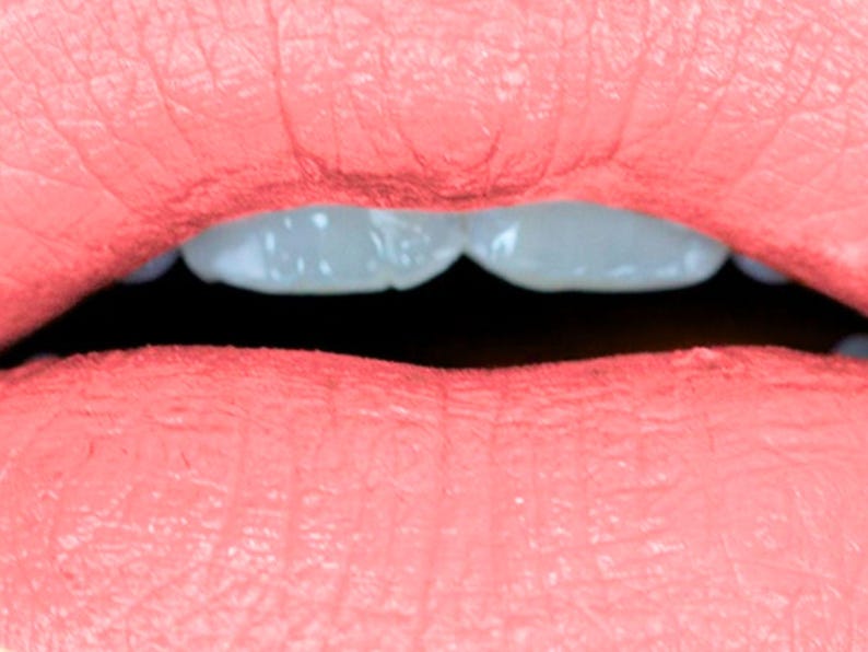 May include: Close-up of a person's lips, painted with a coral-colored lipstick. The lips are slightly parted, revealing a glimpse of white teeth against a dark background. The texture of the lips is visible.