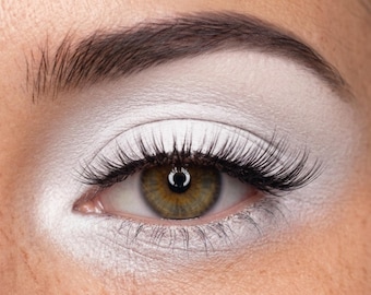 May include: Close-up of an eye with dramatic makeup. The eye has long, black eyelashes and a light green iris. The eyelid and brow are covered in white eyeshadow, and the eyebrow is dark brown.