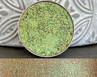 May include: A round, silver-colored makeup palette with a green, glittery eyeshadow inside. The eyeshadow has a shimmery, iridescent finish.