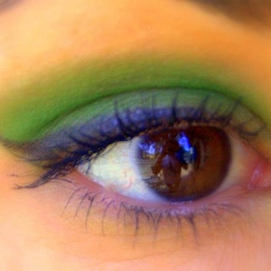 Puede incluir: Primer plano de un ojo con iris marr&oacute;n y largas pesta&ntilde;as. El p&aacute;rpado superior est&aacute; pintado con sombra de ojos verde, y el p&aacute;rpado inferior est&aacute; delineado con delineador de ojos azul oscuro. La piel alrededor del ojo tiene un tono melocot&oacute;n.