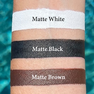 Pode incluir: Um close-up de tr&ecirc;s amostras de tinta fosca no bra&ccedil;o de uma pessoa. As amostras s&atilde;o rotuladas como "Matte White", "Matte Black" e "Matte Brown".