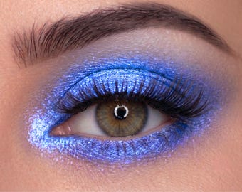 May include: Close-up of an eye with vibrant blue eyeshadow and long, dark eyelashes. The eye has a light brown iris and a defined eyebrow. The makeup creates a dramatic and glamorous look, perfect for beauty and cosmetic products.