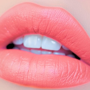 May include: Close-up of a person's lips, painted with a vibrant coral lipstick. The lips are full and well-defined, with a matte finish. The teeth are bright white, contrasting with the coral color.