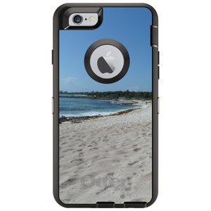 May include: Black OtterBox phone case with a beach scene on the back. The scene features a sandy beach, blue water, and a clear sky. The OtterBox logo is visible in the bottom right corner.
