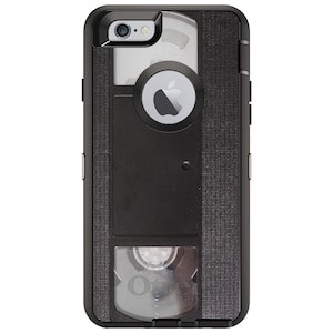 May include: Black OtterBox Defender Series case for an iPhone 6 Plus with a clear, vintage VHS tape design. The case has a textured, black finish and a clear window that shows the Apple logo on the back of the phone.