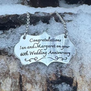May include: Silver pendant with a decorative border and the text "Congratulations Ian and Margaret on your 60th Wedding Anniversary".