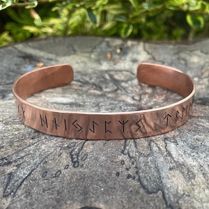 May include: A copper bracelet with a row of Norse runes engraved on the band. The runes spell "NHISJCYYS TRM".