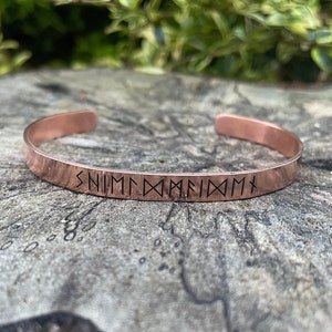 May include: A copper bracelet with a band of runes engraved on the outside. The runes are in black and read "SNIMMM DAAFIIDMMX".