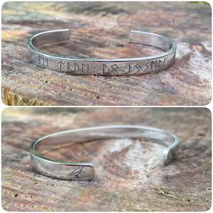May include: A silver bangle bracelet with a bird design on one side and the inscription "BM ↑ TRNM ↑ ᛦ ↑ ᛦSMM" on the other side.