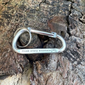 May include: Silver carabiner keychain with the text "Climb every mountain" engraved on it. The keychain has a small mountain logo.