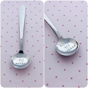 Puede incluir: Una cuchara pequeña de acero inoxidable con un cuenco redondeado. El texto "Daddy's egg BASHER" está estampado en el cuenco. La cuchara se muestra sobre un fondo rosa con lunares blancos.