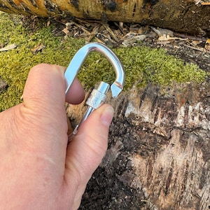 May include: A silver carabiner with a gate that is open. The carabiner is held in a hand.