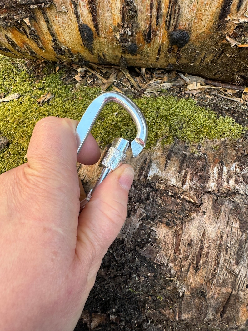 May include: A silver carabiner with a gate that is open. The carabiner is held in a hand.