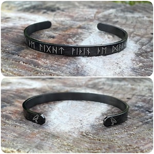 May include: A black metal bangle bracelet with a Norse rune inscription and a small bird design on each end.